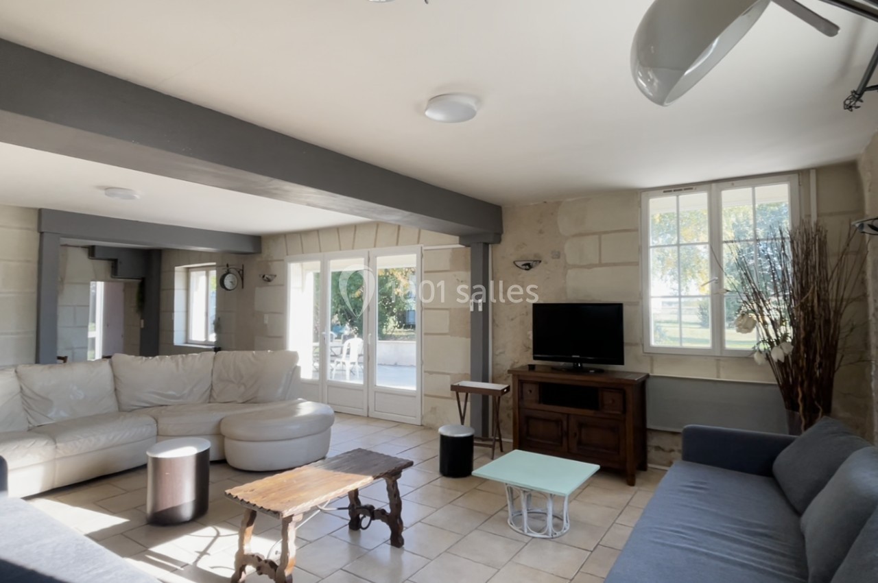 Salon lumineux avec canapés, table basse en bois, télévision sur meuble ancien et grandes fenêtres donnant sur un jardin.