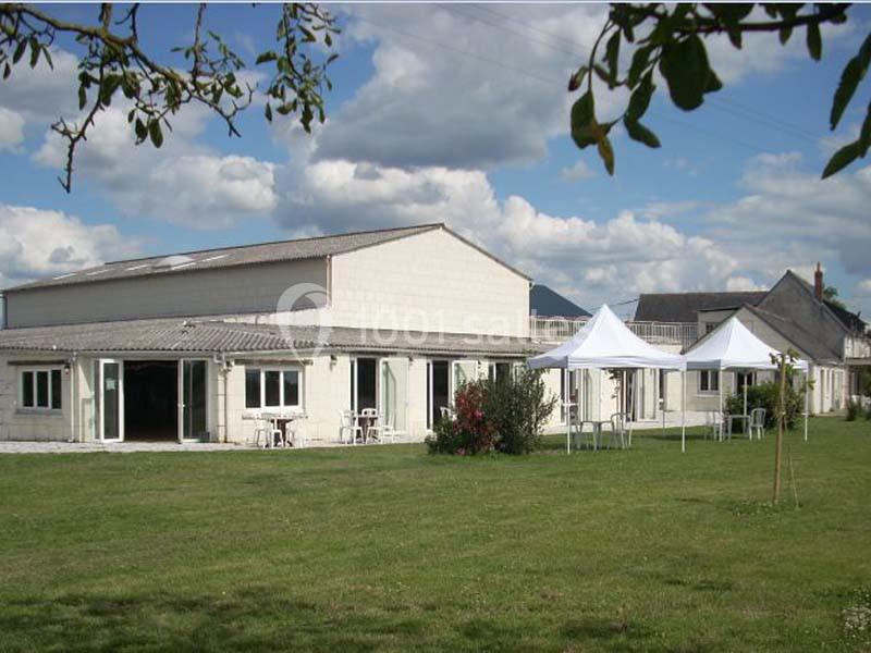 Bâtiment clair avec de grandes fenêtres, entouré d'une pelouse et de tentes blanches sous un ciel partiellement nuageux.