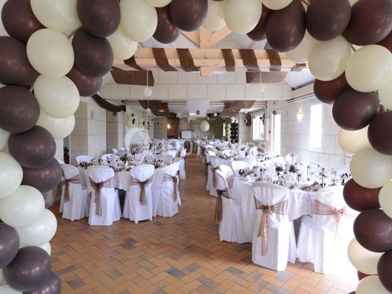 Salle de réception décorée avec des tables dressées et des chaises blanches, encadrée par une arche de ballons.