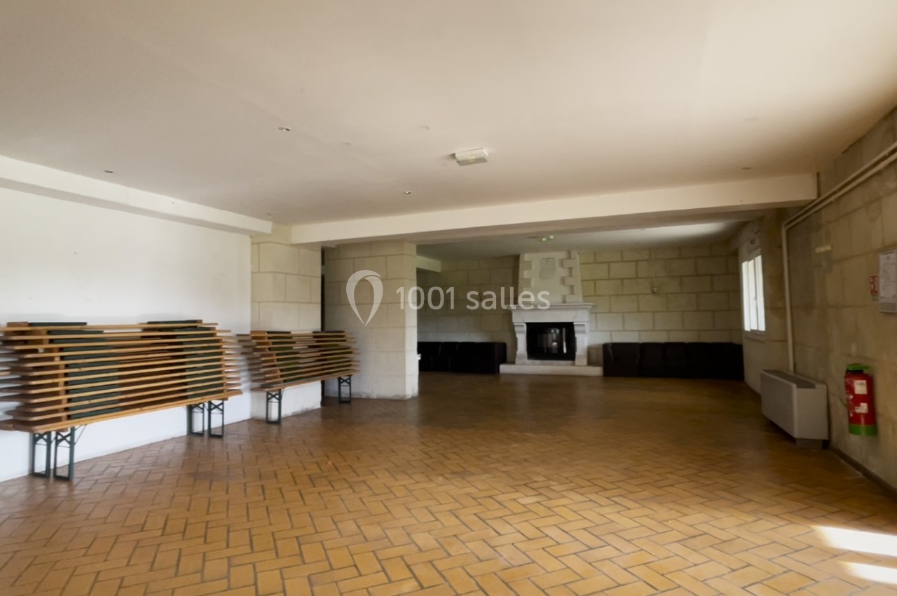 Salle vide avec sol en carrelage, bancs empilés sur la gauche, cheminée au fond et murs en pierre.