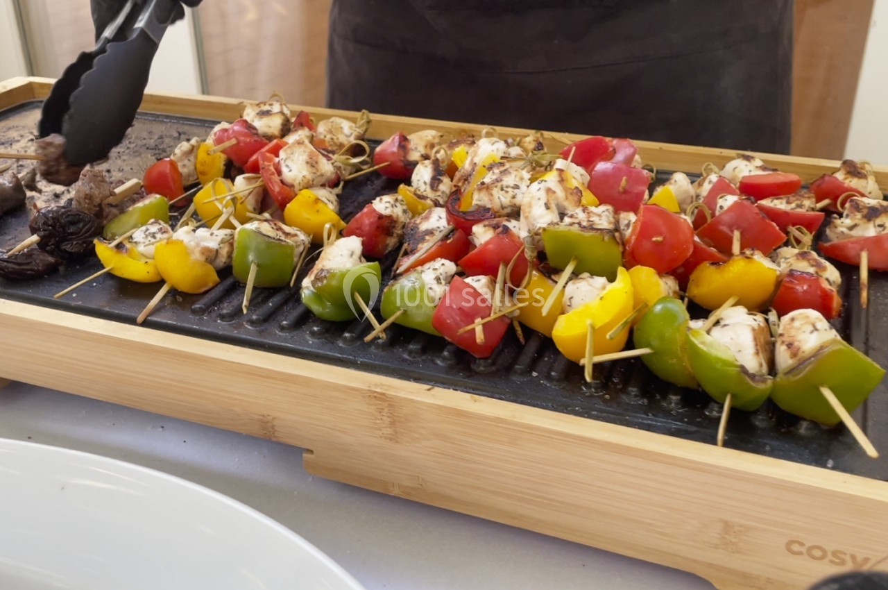Brochettes de légumes et de poulet grillées sur une plancha en bois, avec des pinces tenant un morceau.