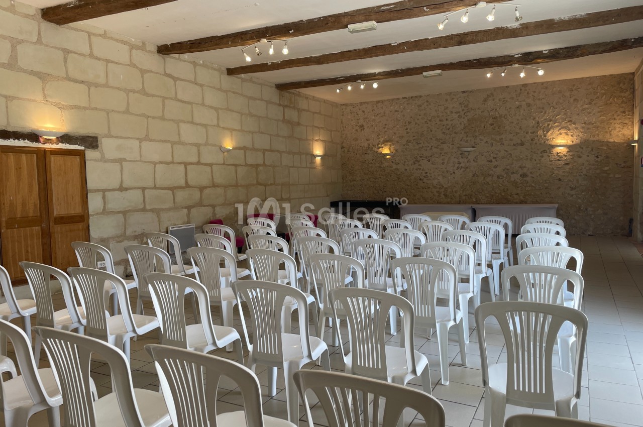 Salle en pierre avec des rangées de chaises blanches en plastique disposées face à une table et un piano.