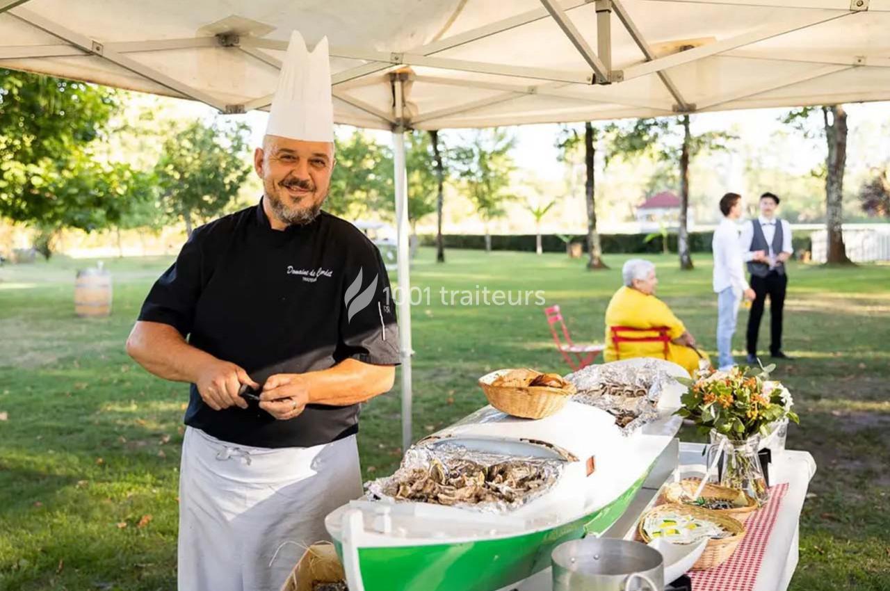 Traiteur Arsac (Gironde) - Domaine de Cordet #8 Un chef en tenue prépare des huîtres sous une tente dans un cadre extérieur verdoyant avec des invités en arrière-plan.