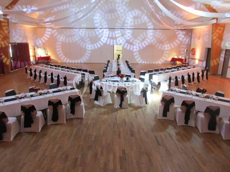 Salle de réception décorée avec des tables dressées en U, nappes blanches et nœuds noirs sur les chaises.