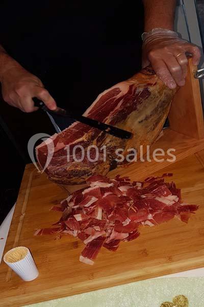 Une personne découpe finement un jambon sec sur une planche en bois, avec des tranches déjà empilées au premier plan.