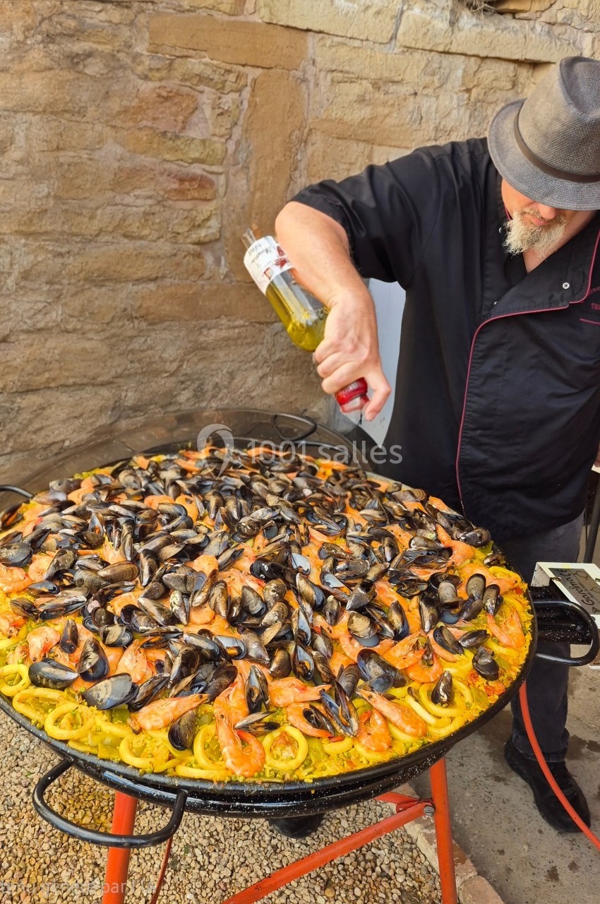 Un cuisinier prépare une grande paella garnie de moules, crevettes et calamars dans une poêle extérieure.