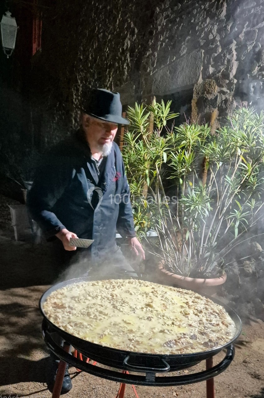 Un homme en chapeau noir cuisine une grande poêle de plat fumant en extérieur, près de plantes et d'un mur en pierre.