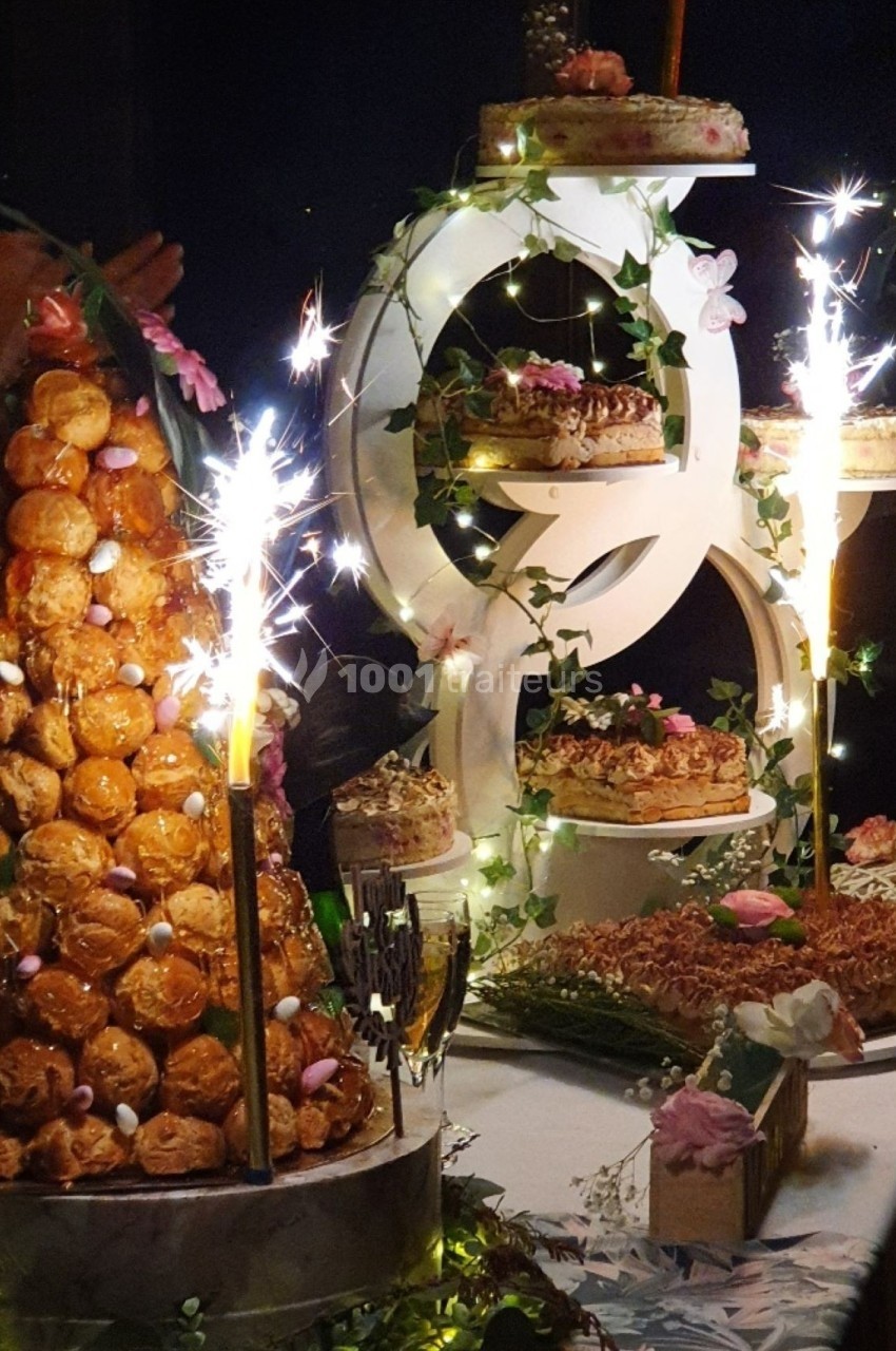 Pièce montée en choux et gâteaux décorés, entourés de guirlandes lumineuses et de fleurs, avec des cierges étincelants.