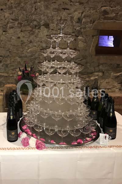 Tour de coupes à champagne empilées sur une table décorée de fleurs, entourée de bouteilles dans un cadre rustique.