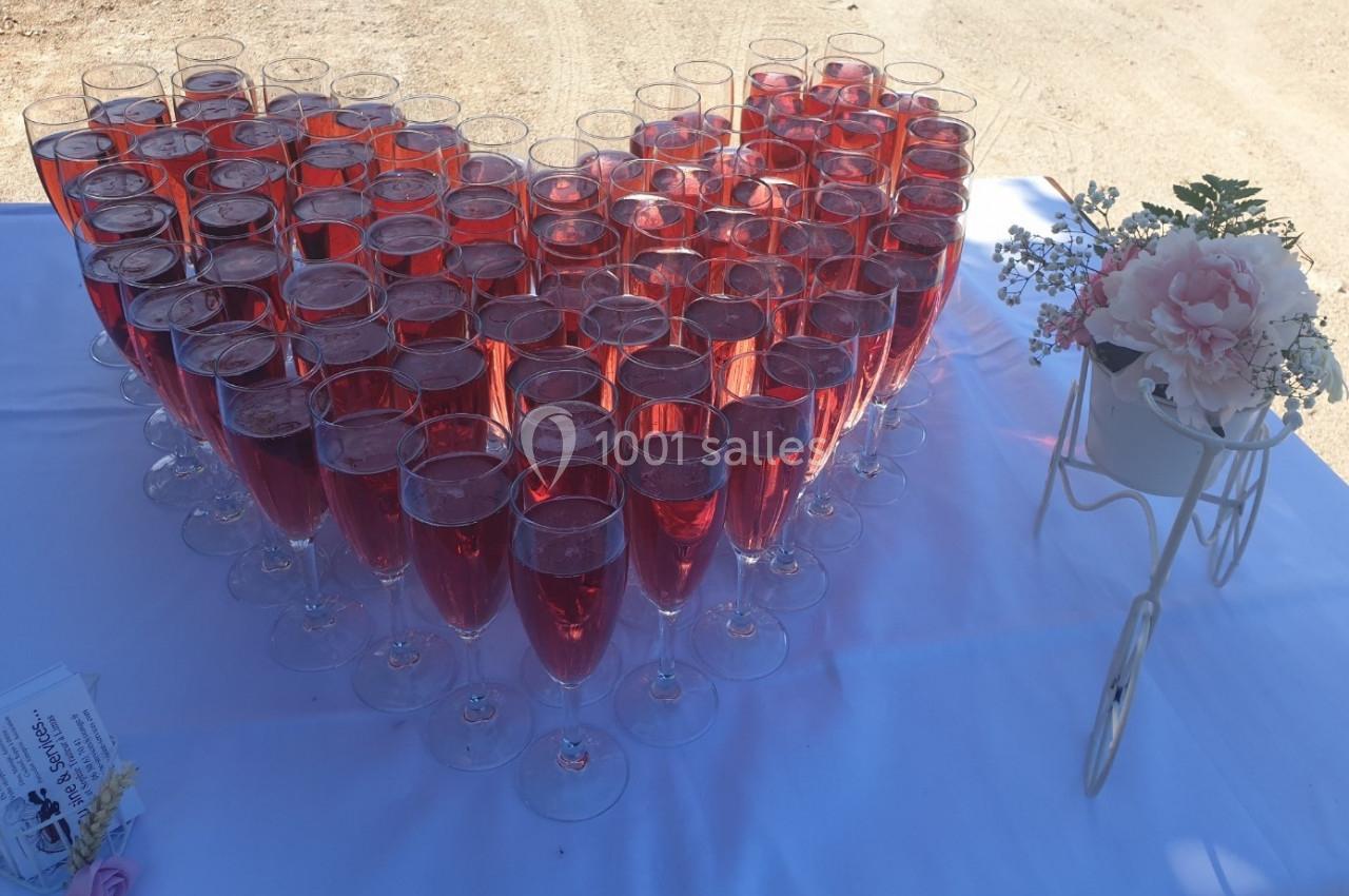 Verres de cocktail rosé disposés en forme de cœur sur une table blanche, avec une décoration florale à côté.