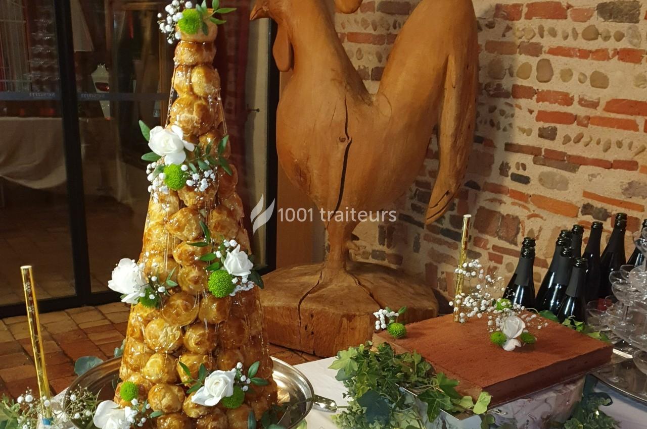 Pièce montée en choux décorée de fleurs, posée sur une table avec un coq sculpté en arrière-plan.