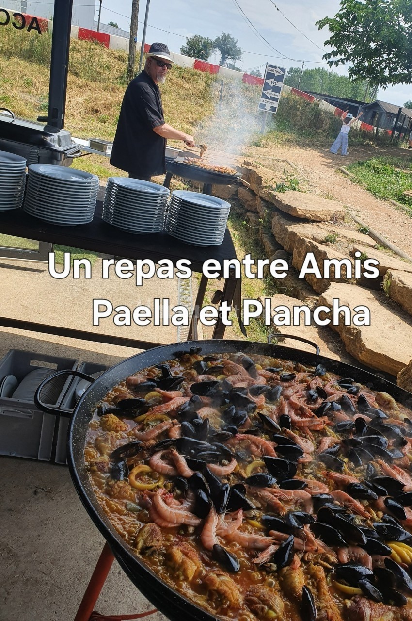 Un cuisinier prépare un repas en plein air avec une grande paella garnie de fruits de mer au premier plan.