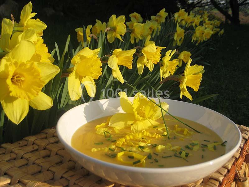 Bol de soupe jaune garni de ciboulette et de fleurs, posé sur une nappe en osier devant un parterre de jonquilles.