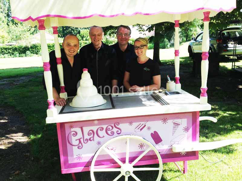 Quatre personnes posent derrière un chariot rose décoré pour la vente de glaces, dans un parc ensoleillé.