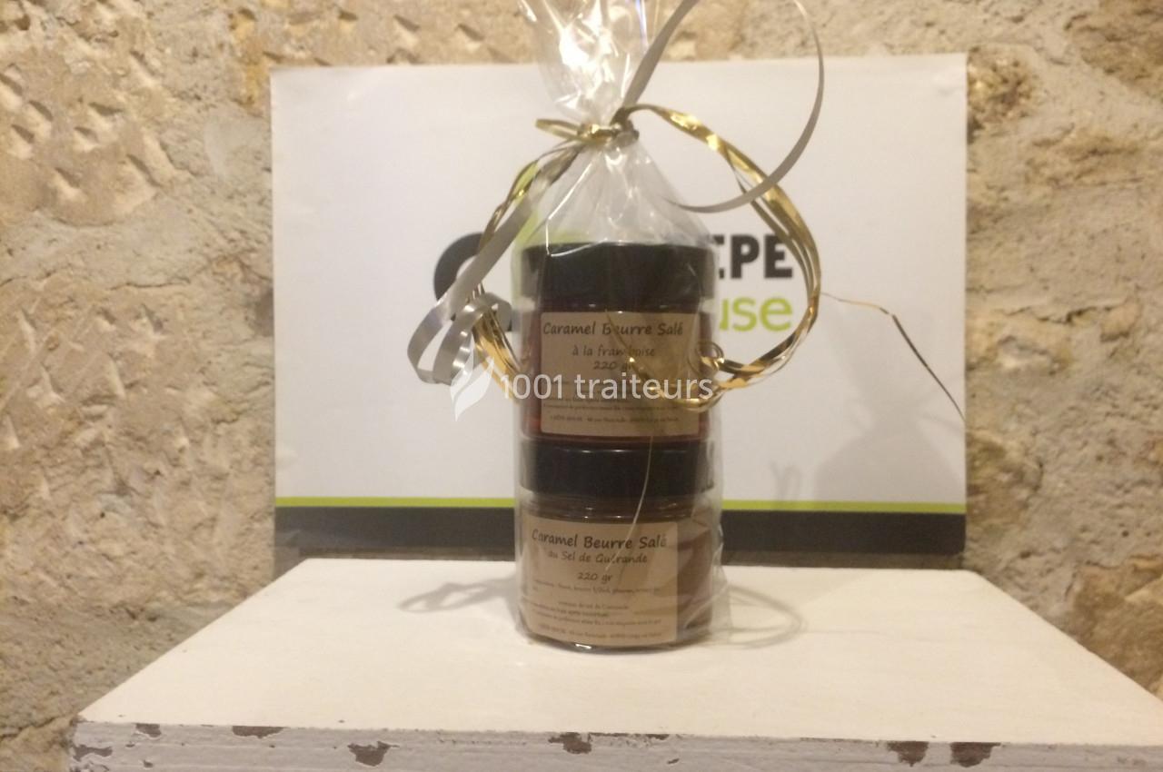 Deux pots de caramel au beurre salé emballés avec un ruban doré, posés sur une surface blanche devant un mur en pierre.