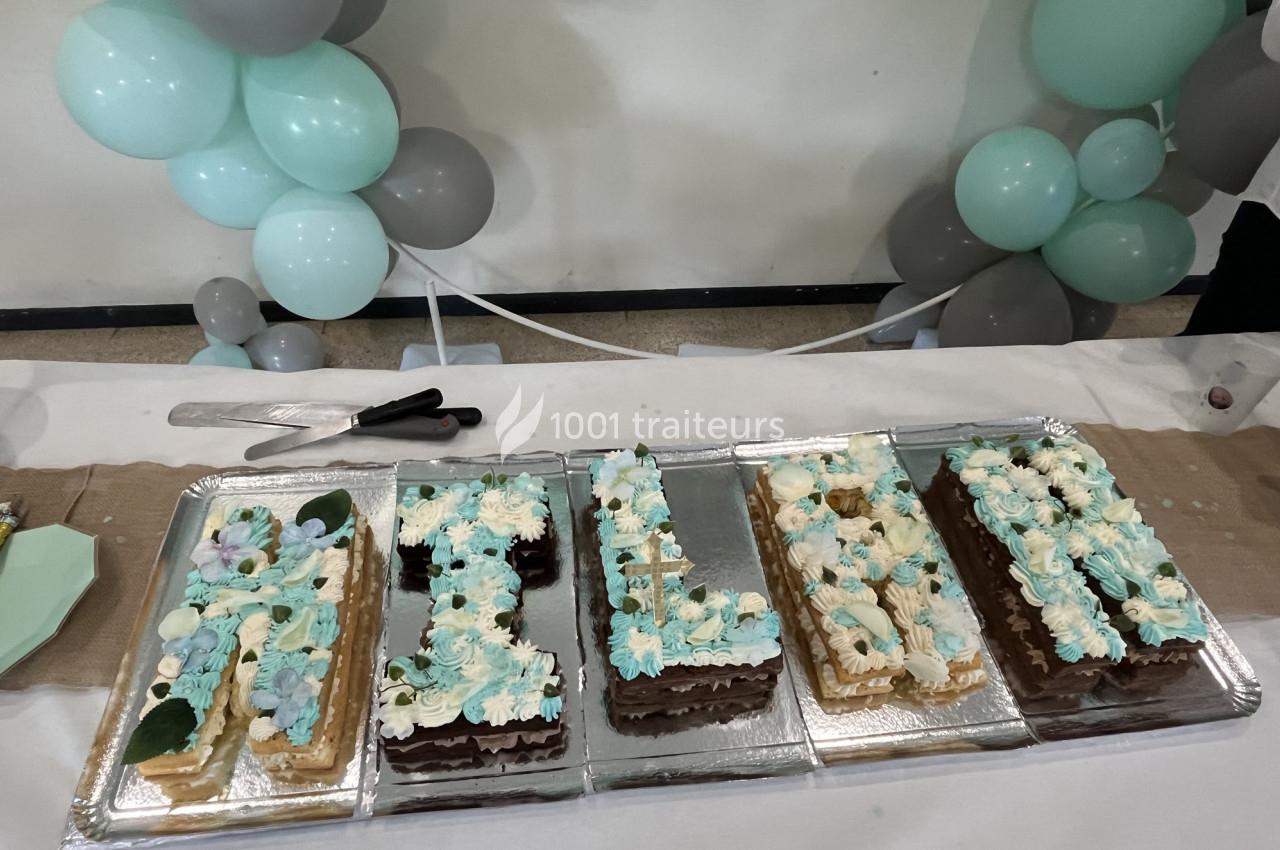 Gâteaux en forme de lettres décorés de crème et fleurs, entourés de ballons verts et gris sur une table blanche.