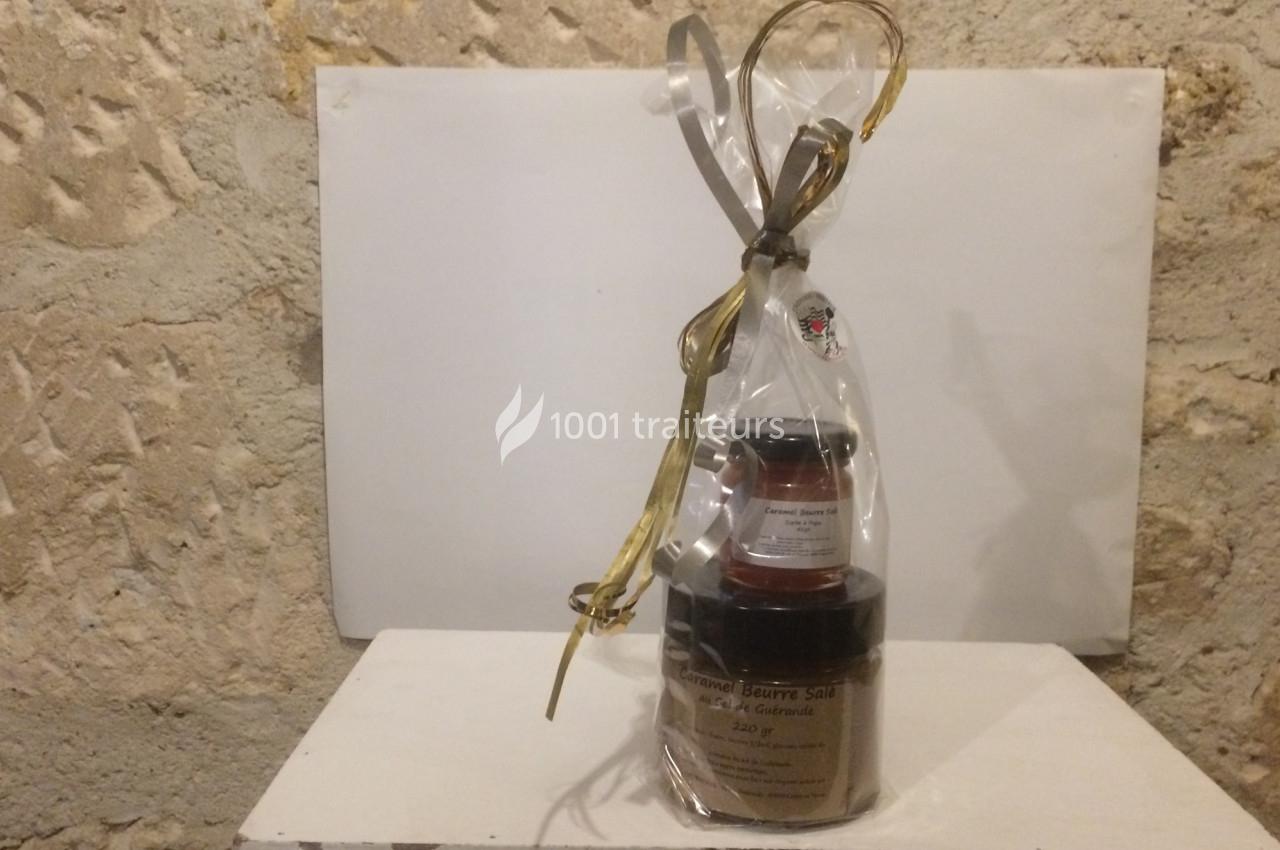 Ensemble de pots de confiture et beurre salé emballés dans un sachet transparent avec ruban doré, posé sur une table blanche.