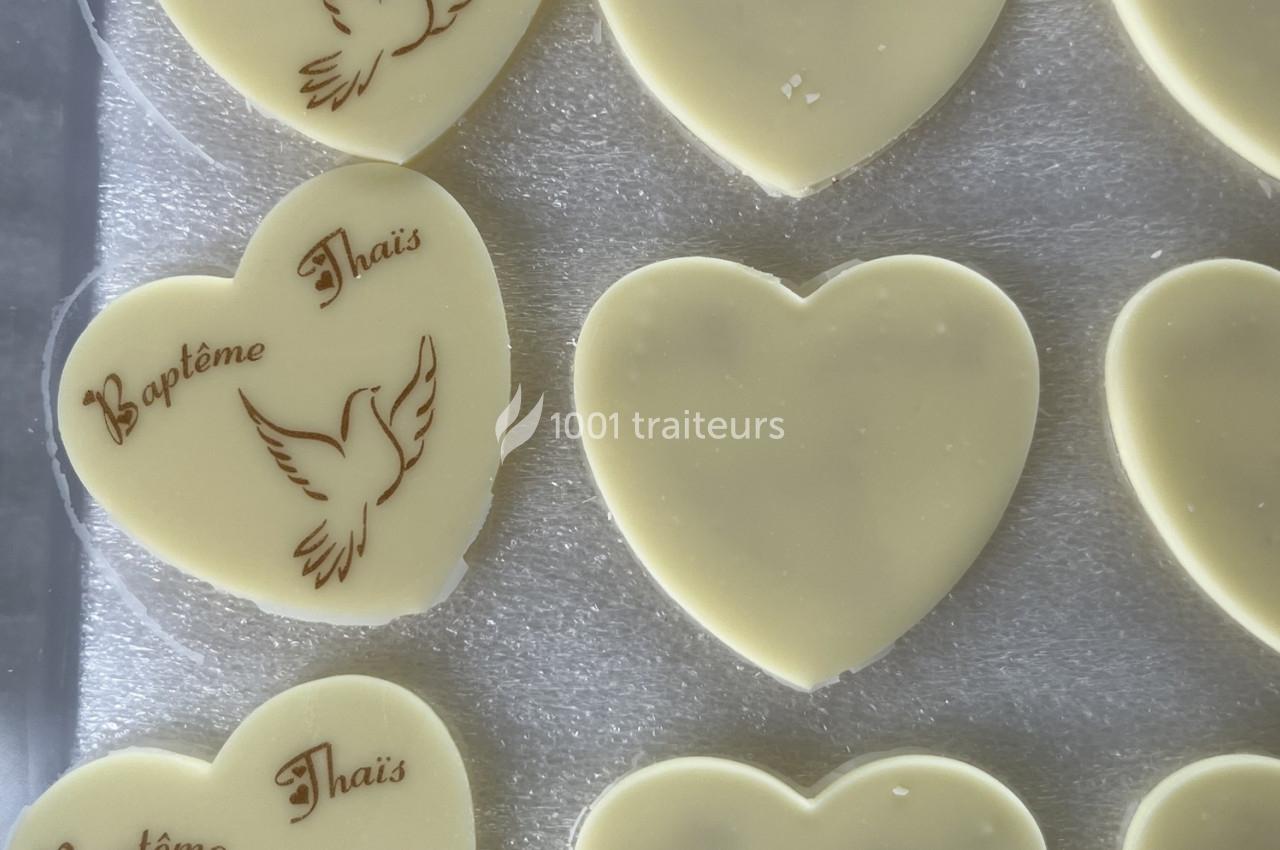 Cœurs en chocolat blanc, certains gravés d'une colombe et du texte ’Baptême Thaïs’, posés sur une surface argentée.