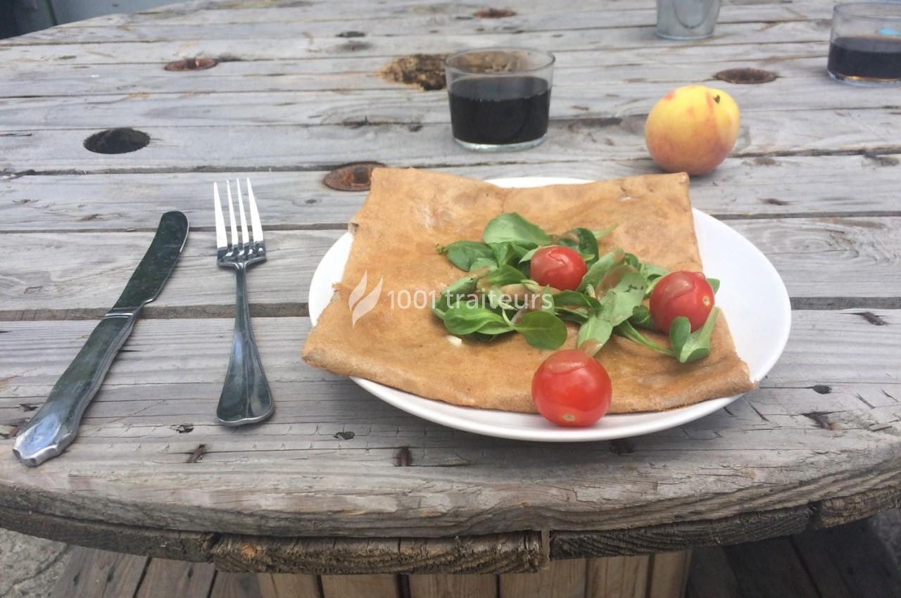 Assiette avec une galette garnie de mâche et tomates cerises, posée sur une table en bois avec des couverts et un verre.