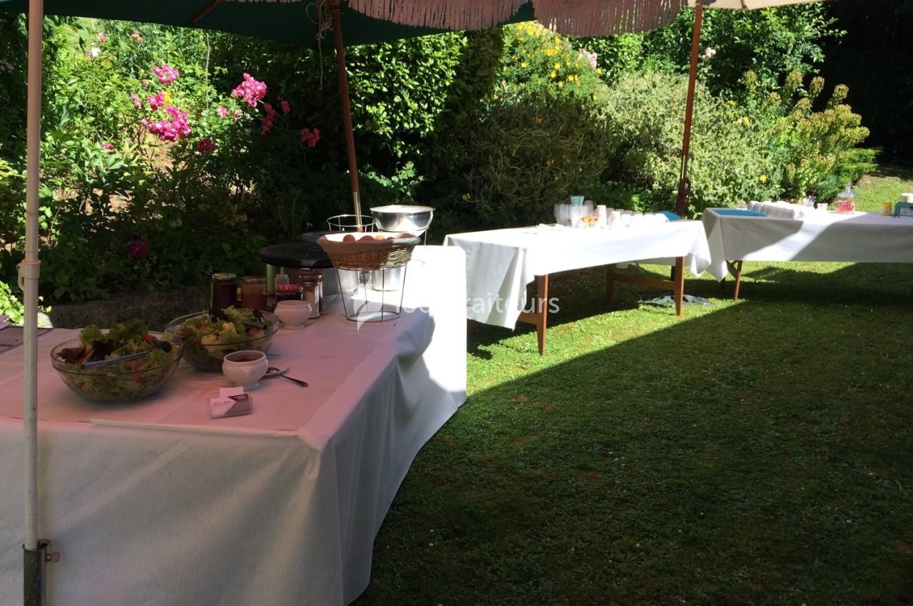 Tables dressées sous des parasols dans un jardin, avec salades, plats et vaisselle pour un buffet en plein air.