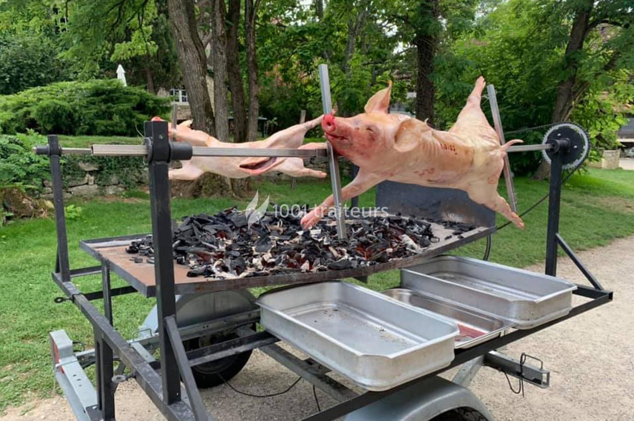 Cochon entier en cours de cuisson sur une broche rotative au-dessus de braises, dans un cadre extérieur verdoyant.
