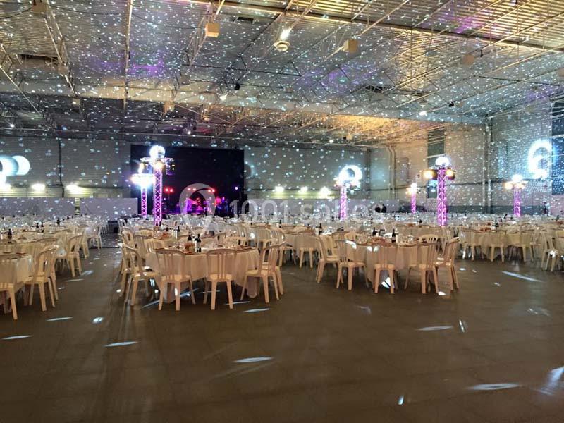 Salle de réception décorée avec des tables rondes, des chaises blanches et des éclairages lumineux projetant des motifs.