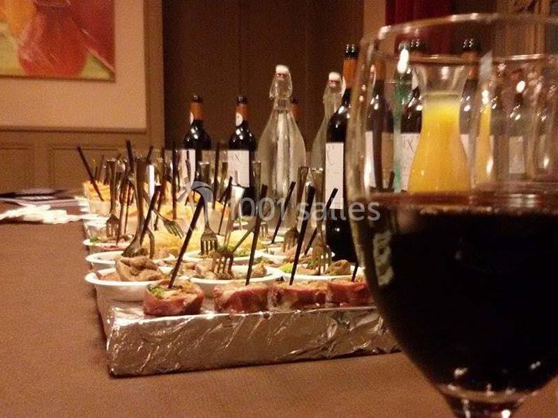 Table garnie de plats variés, bouteilles de vin et jus, avec un verre de vin rouge au premier plan.