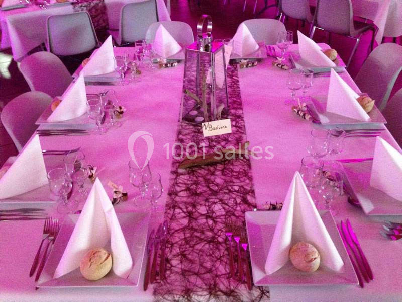 Table dressée pour un événement, avec nappes blanches, serviettes pliées, couverts, verres et décoration violette.