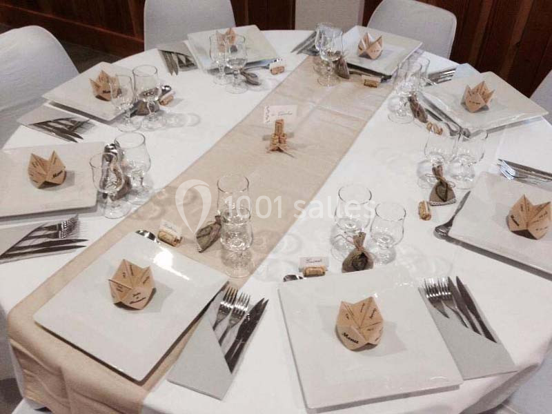 Table dressée avec une nappe blanche, chemin de table beige, assiettes carrées, verres et pliages en papier décoratifs.