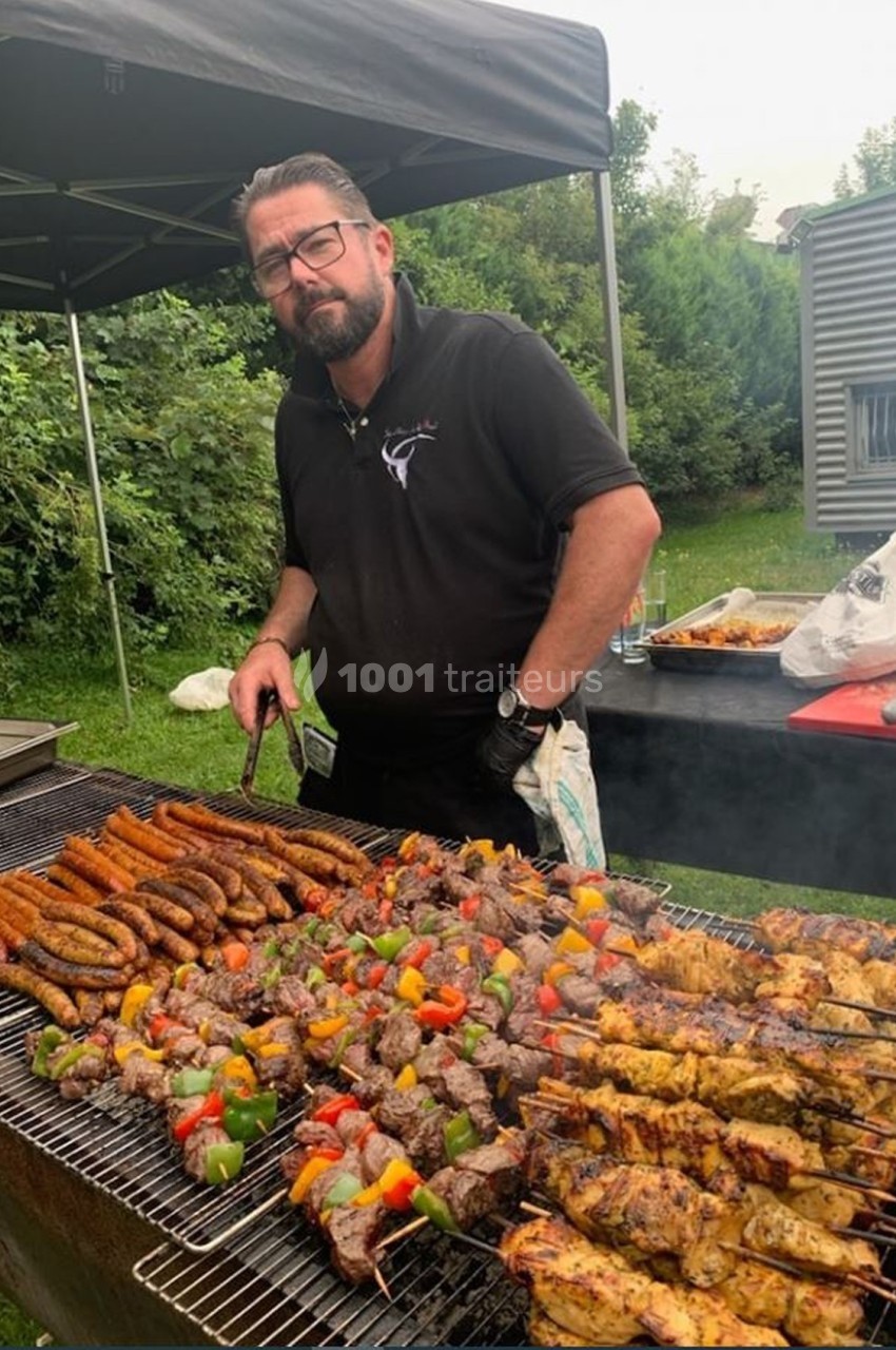 Homme cuisinant des brochettes et saucisses sur un barbecue en plein air, entouré de verdure et d'équipements.