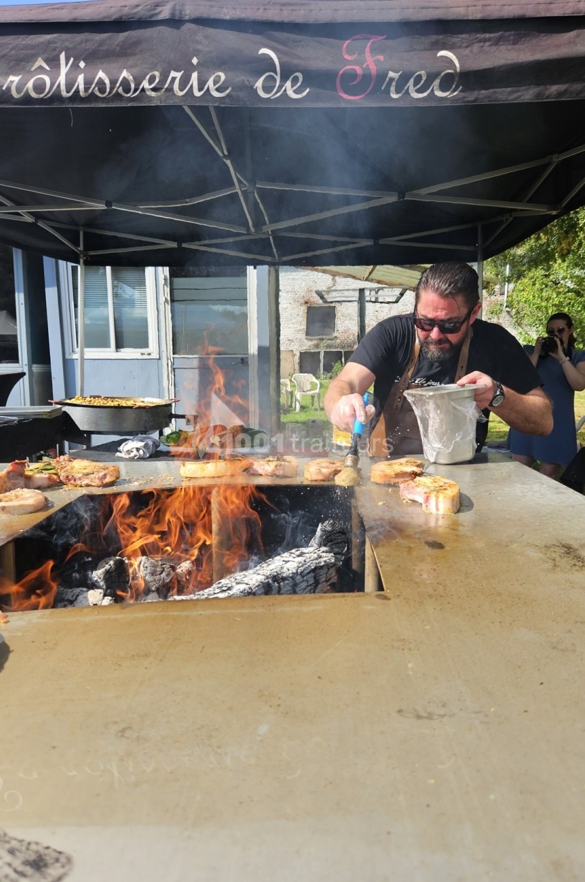 Un cuisinier prépare des plats au feu de bois sous un stand, avec des flammes vives et des aliments en cuisson.
