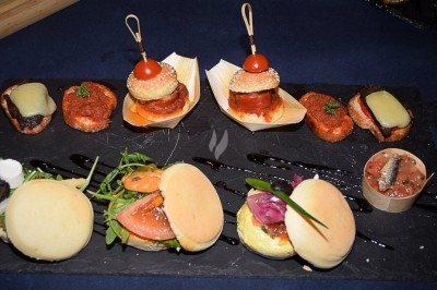 Plateau de mini-burgers, bouchées variées et garnitures sur une ardoise noire, présenté sur une nappe sombre.