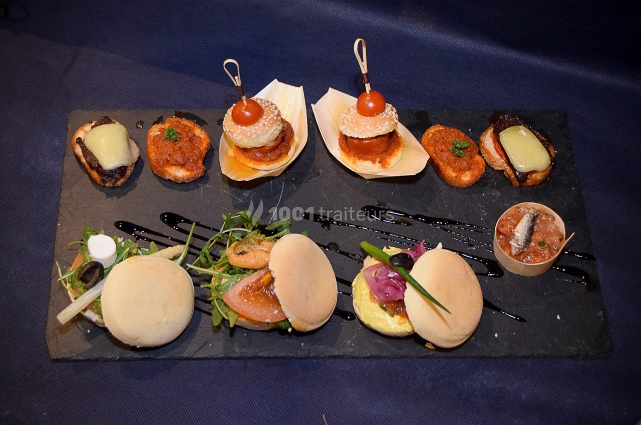 Plateau de mini-burgers, bouchées variées et garnitures sur une ardoise noire, présenté sur une nappe sombre.