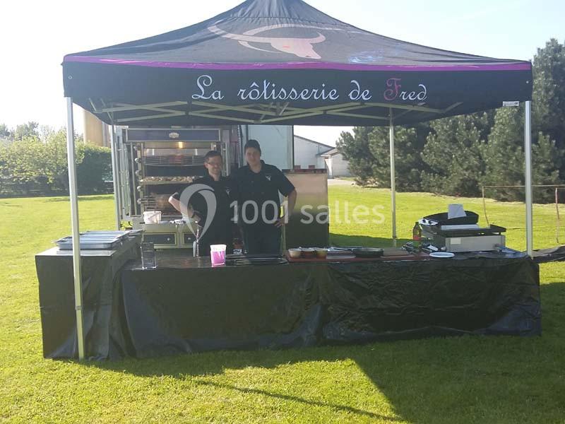 Deux personnes sous une tente de rôtisserie installée en extérieur, avec équipements de cuisine visibles.