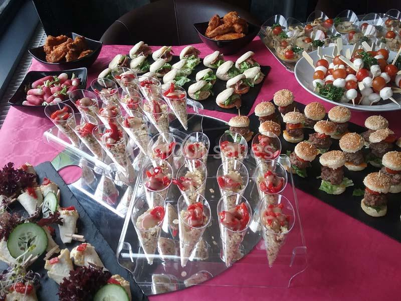 Buffet avec mini-burgers, verrines, brochettes apéritives, sandwichs et légumes sur une table rose.