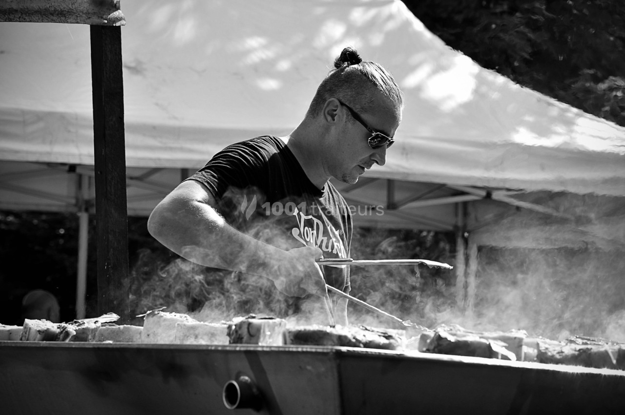 Homme cuisinant des aliments sur un barbecue en plein air, entouré de fumée et d'un chapiteau en arrière-plan.