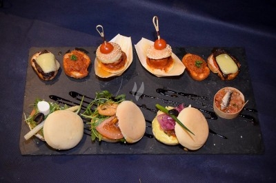Plateau de mini-burgers, bouchées variées et garnitures sur une ardoise noire, présenté sur une nappe sombre.