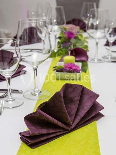 Table dressée avec serviettes pliées violettes, chemin de table vert, verres et décoration florale au centre.