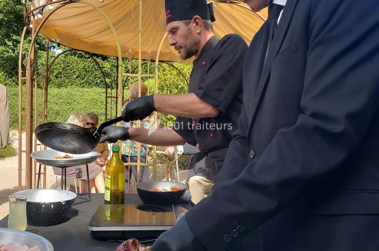 Un chef en tenue noire cuisine en plein air sous une tonnelle, assisté par un homme en costume.