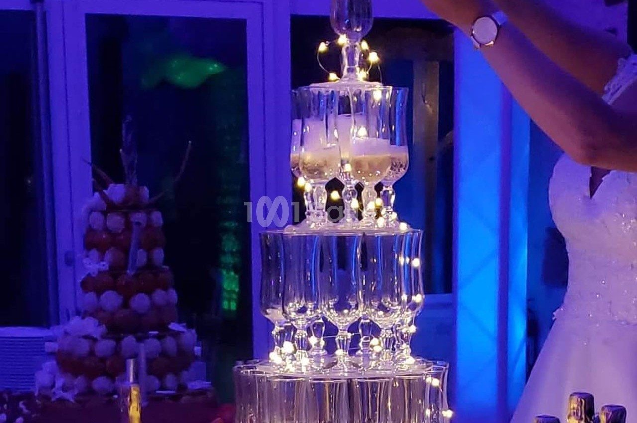 Tour de coupes de champagne illuminée, surmontée d'une coupe remplie, dans un décor de réception festive.