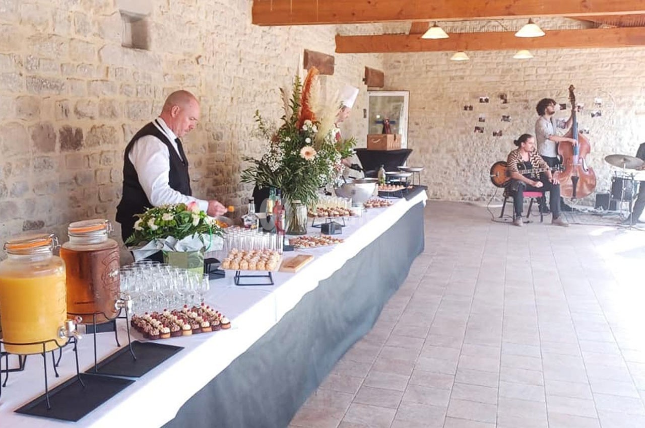 Buffet dressé dans une salle en pierre avec un serveur, des boissons, des plats et un duo musical en arrière-plan.