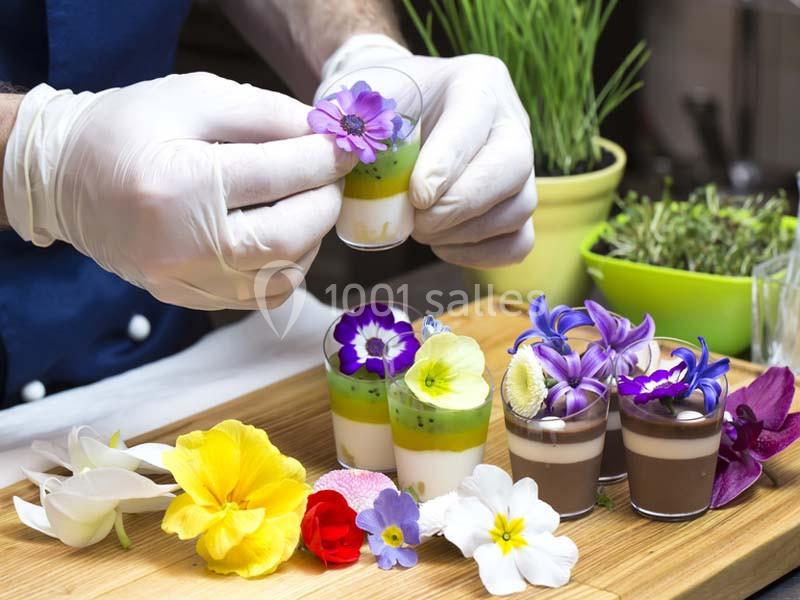 Un chef décore des verrines sucrées avec des fleurs comestibles sur une planche en bois, entourée d'ingrédients frais.