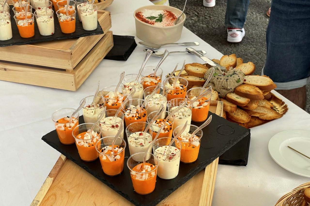 Verrines de soupe froide orange et blanche garnies d'herbes, servies sur un plateau avec des toasts et des sauces.