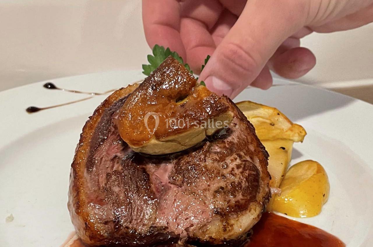 Un morceau de viande saignant garni de foie gras, accompagné de pommes de terre et d'une sauce, sur une assiette blanche.