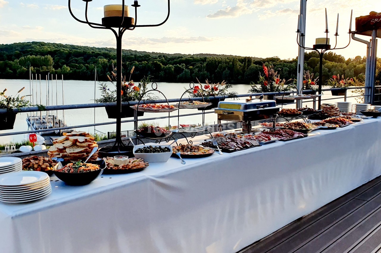 Buffet en plein air avec divers plats et assiettes, installé sur une terrasse surplombant un lac au coucher du soleil.