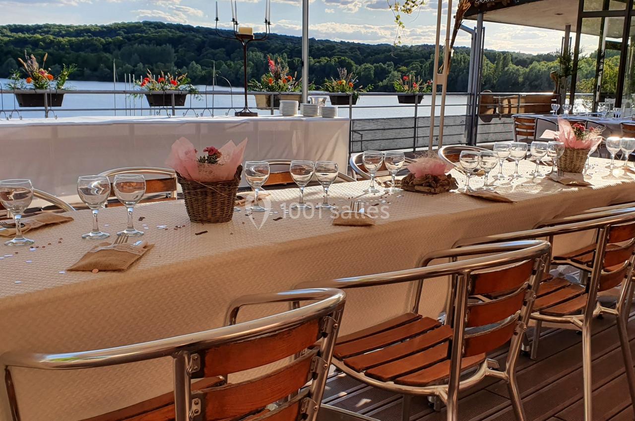 Table dressée avec nappes beiges, vaisselle élégante et décorations florales, sur une terrasse avec vue sur un lac et forêt.