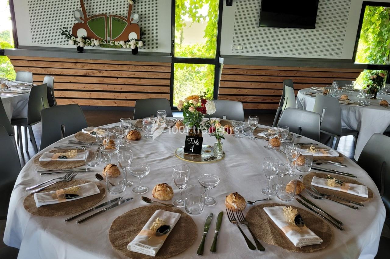 Table ronde dressée pour un repas, avec nappes blanches, couverts, verres, fleurs et décoration de salle lumineuse.