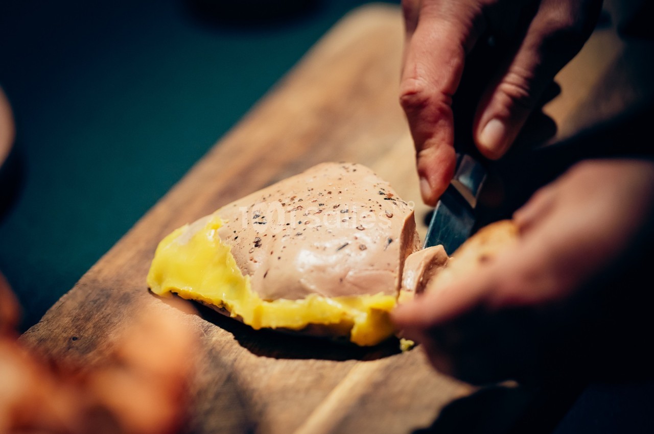 Une main coupe une tranche de foie gras posé sur une planche en bois, accompagné d'une garniture jaune.