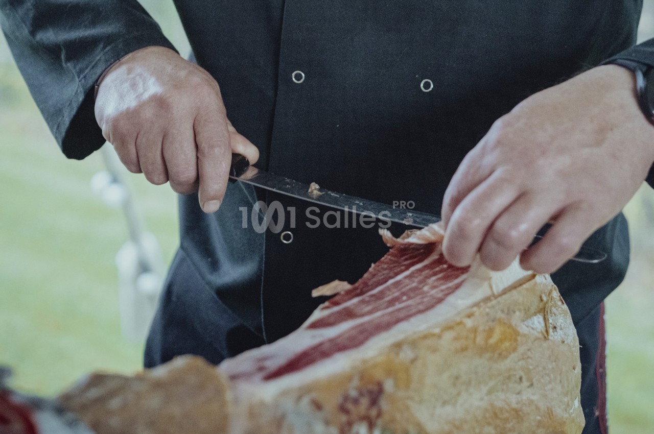 Un cuisinier découpe finement une tranche de jambon cru à l'aide d'un couteau.