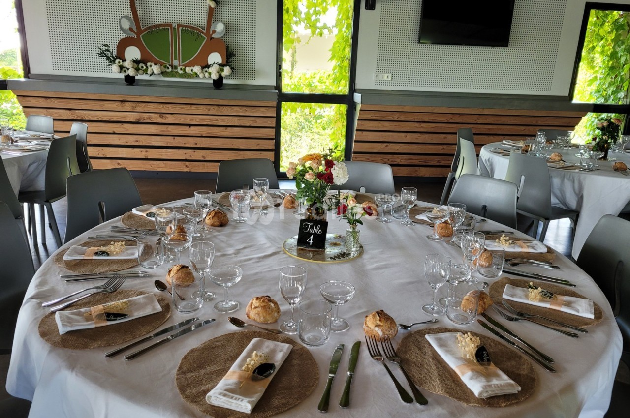 Table ronde dressée pour un repas, avec nappes blanches, couverts, verres, fleurs et décoration de salle lumineuse.